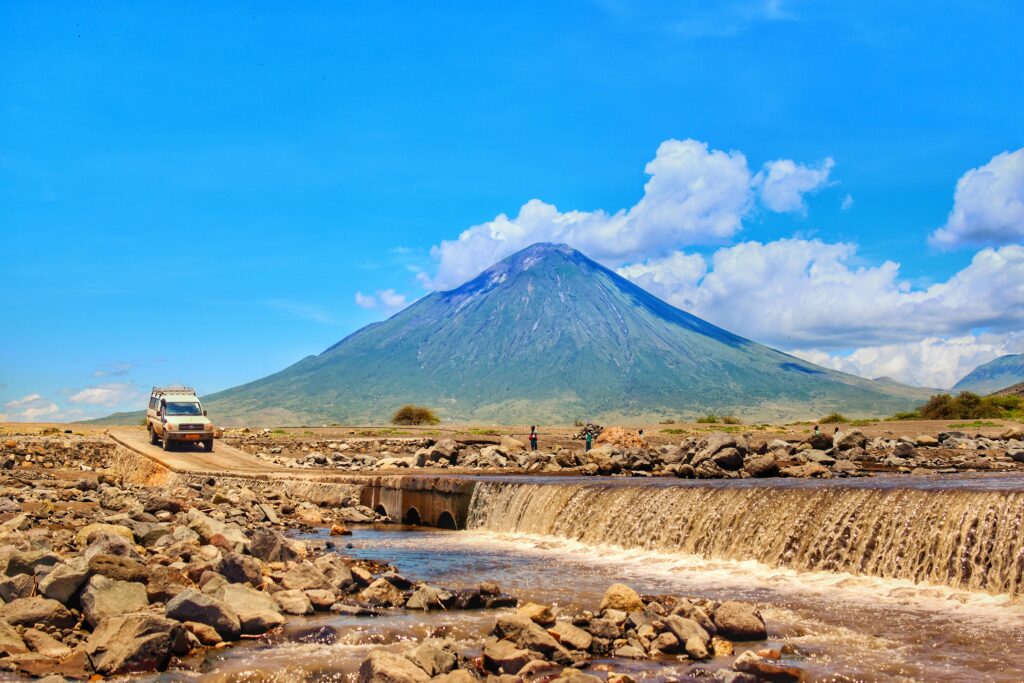 A rugged vehicle crosses a rocky riverbed with the majestic Mount Meru in the background, showcasing Tanzania's wild landscapes.