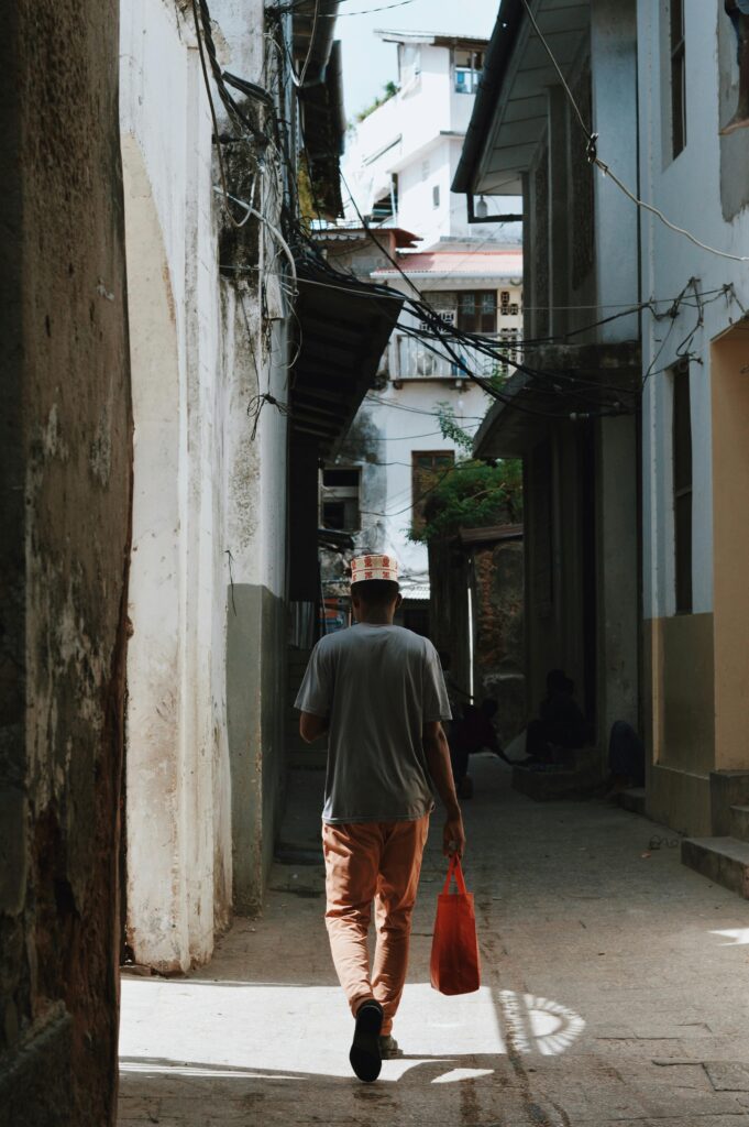 A man walks through the narrow, sunlit streets of Zanzibar City, carrying a red bag.